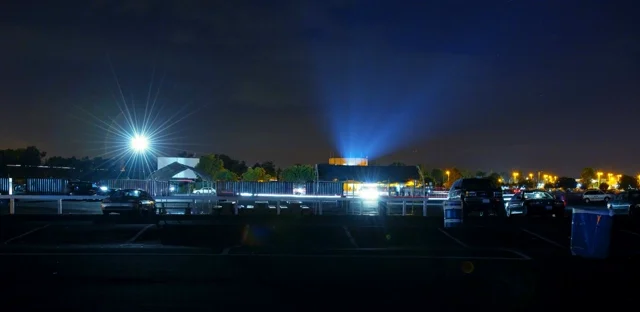 A packed drive-in movie theater at night, rows of cars facing a large illuminated screen